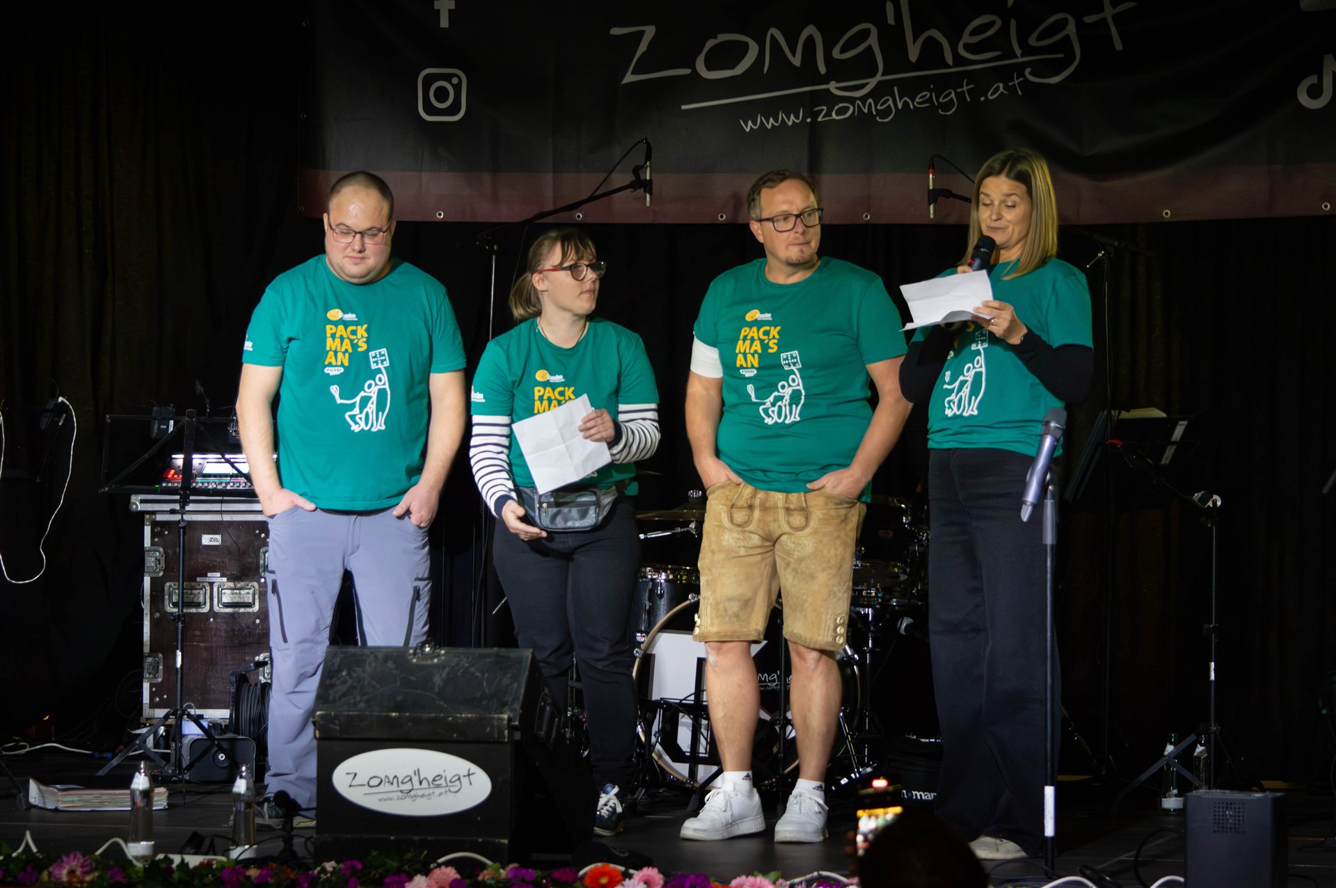 Vier Moderierende in Pack-ma's-an-Shirts auf der Bühne