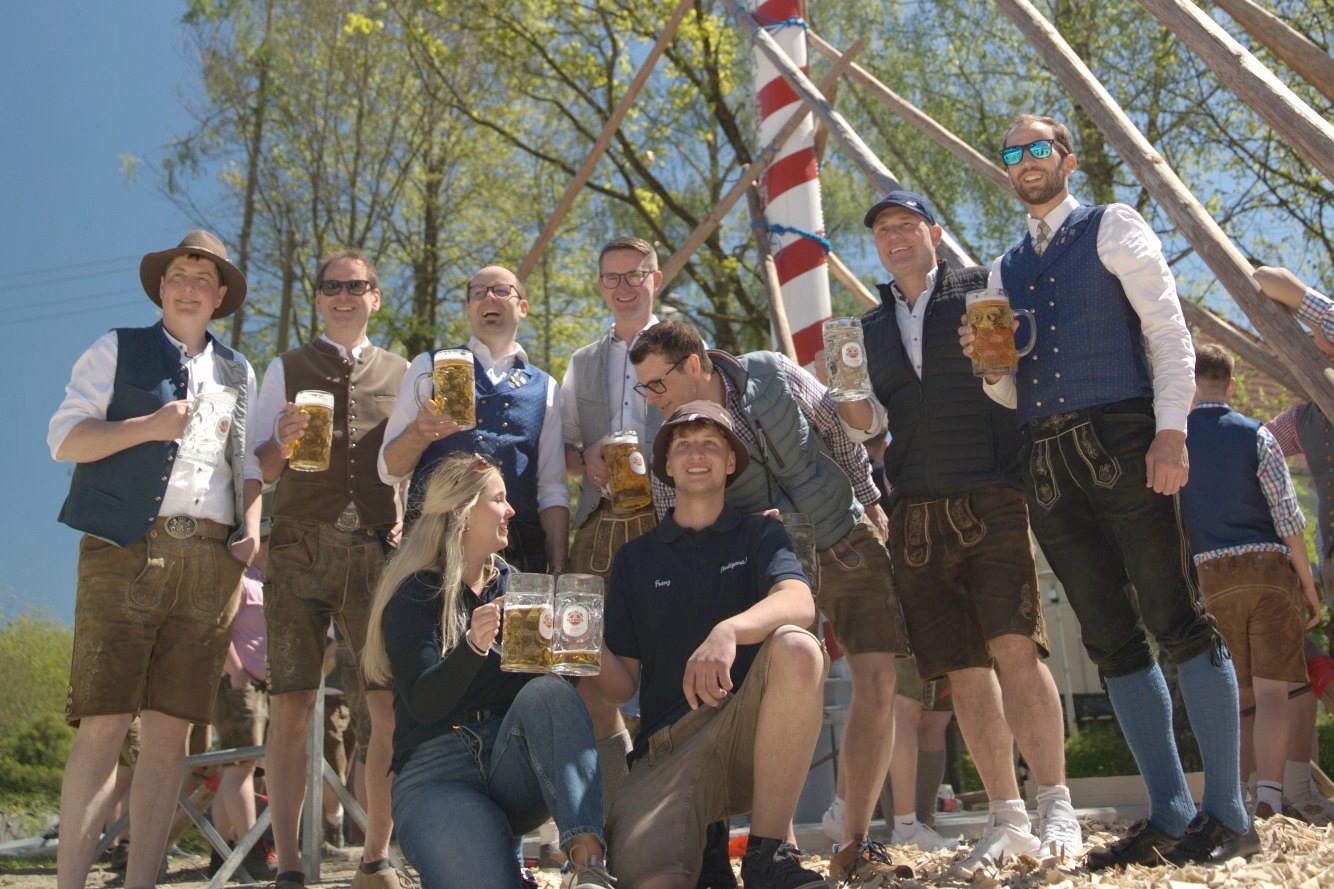 Gruppenfoto in Tracht mit Bierkrügen unter dem aufgestellten Maibaum